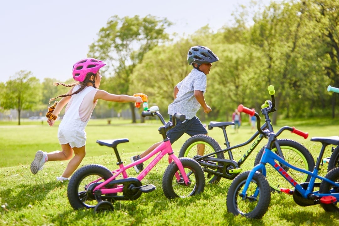Kinder mit Helmen laufen über eine Wiese mit mehreren Kinderfahrrädern.