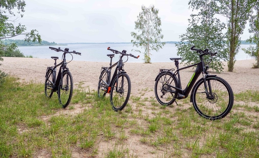 Drei KTM E-Trekkingbikes stehen nebeneinander am Ufer eines Sees.