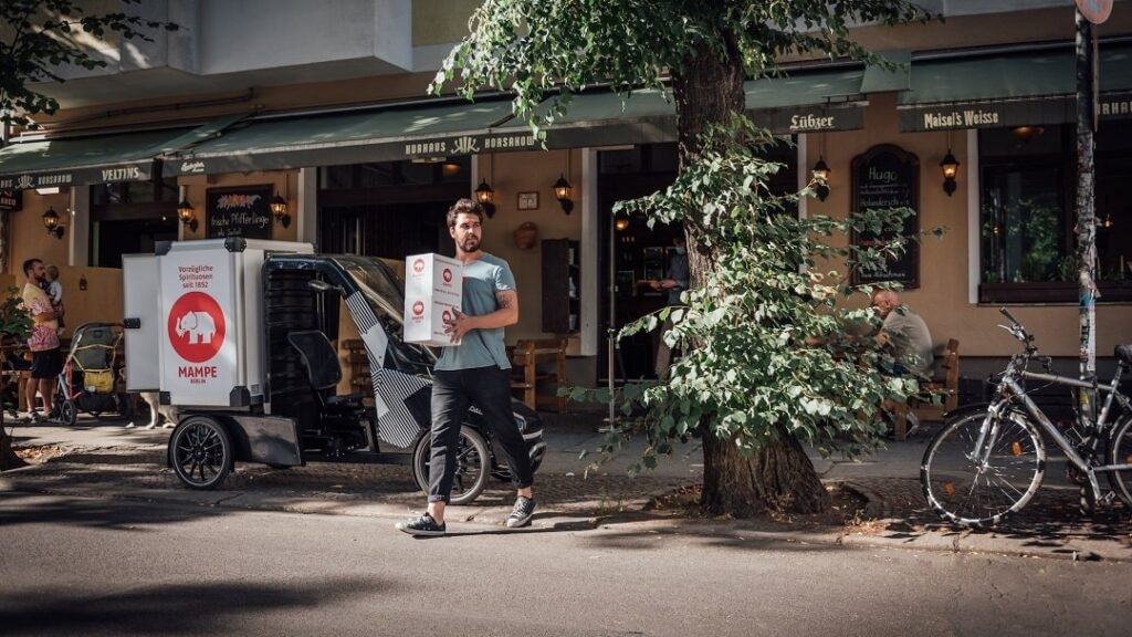 Mann läuft mit einem Paket neben einem Lastenrad vor einem Café über die Straße.