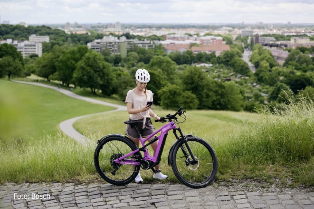Frau mit Fahrradhelm steht neben einem E-Mountainbike und schaut auf ihr Smartphone.