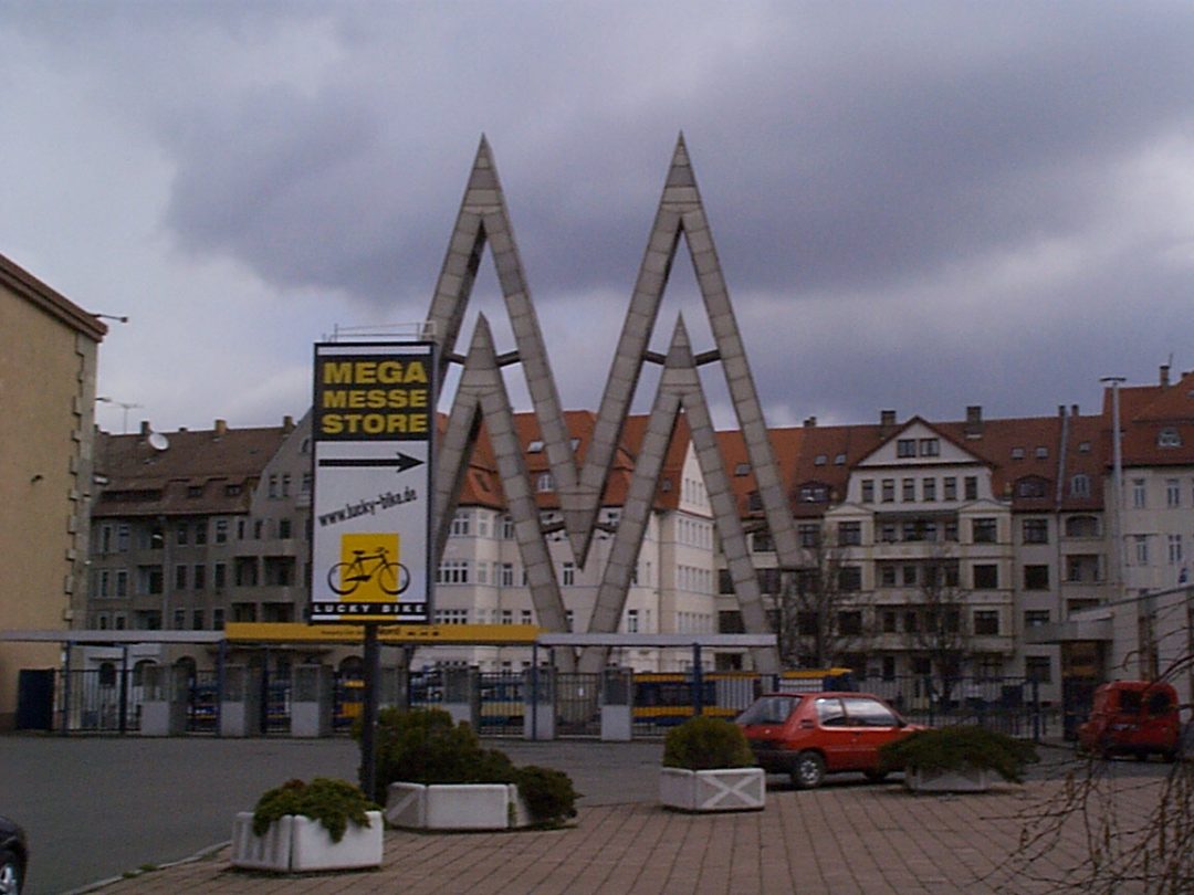 Außenansicht eines Lucky Bike Standorts mit großem „Mega Messe Store“-Schild und markanter Metallkonstruktion vor Wohnhäusern.