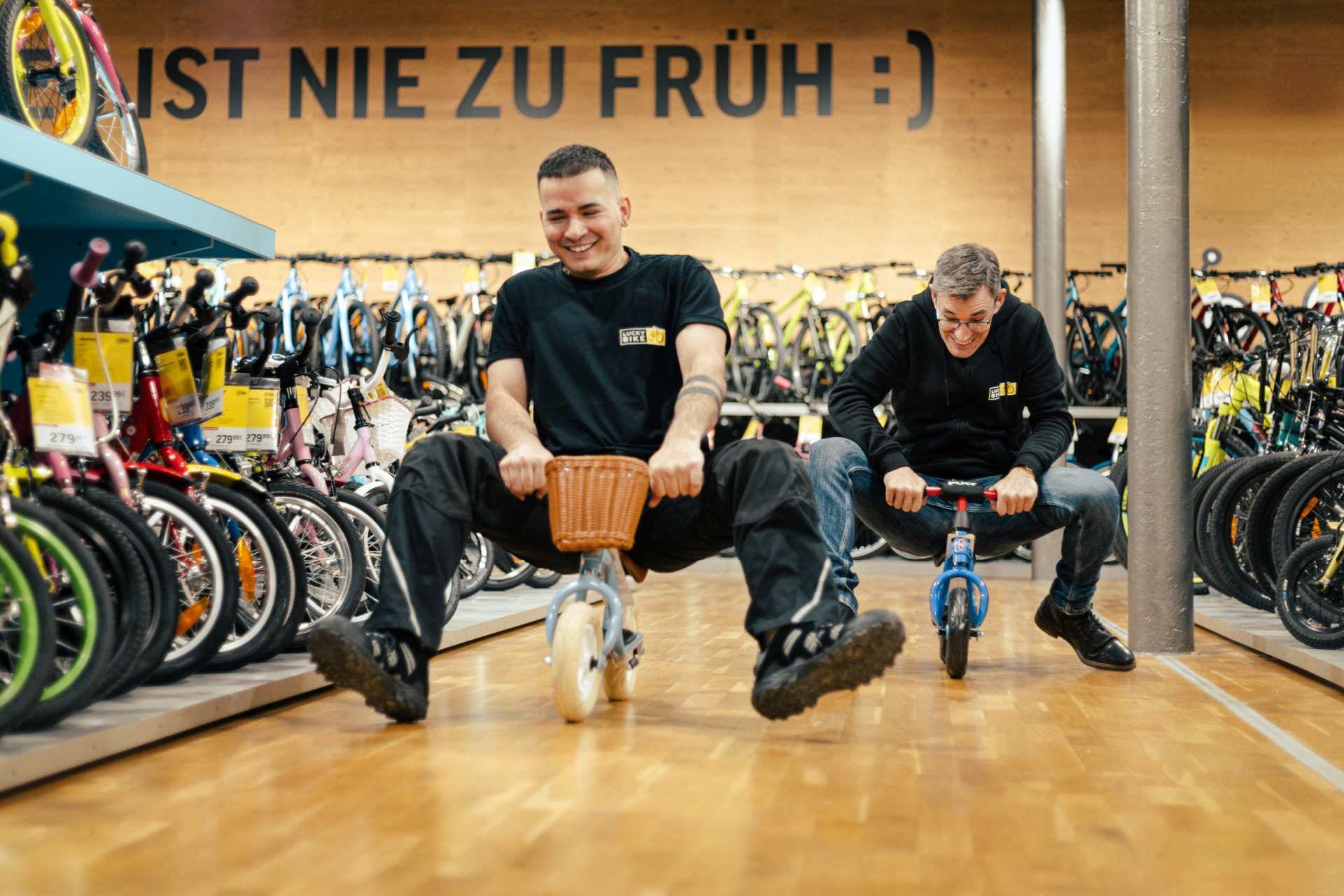 Lucky Bike Mitarbeiter sitzen lachend auf einem Kinderlaufrad.