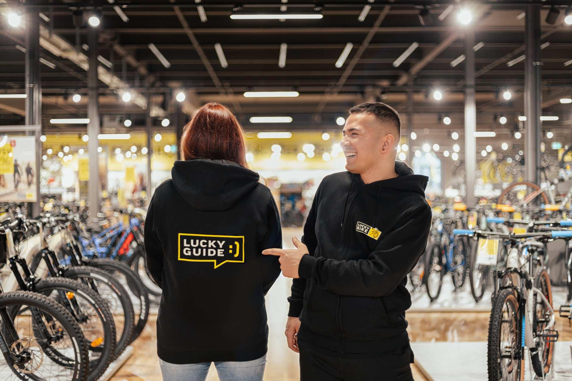 Lucky Bike Mitarbeiter steht lächelnd in einer Fahrradfiliale vor mehreren Fahrrädern und zeigt auf den Hoodie einer Kollegin, auf dem steht: "Lucky Guide".