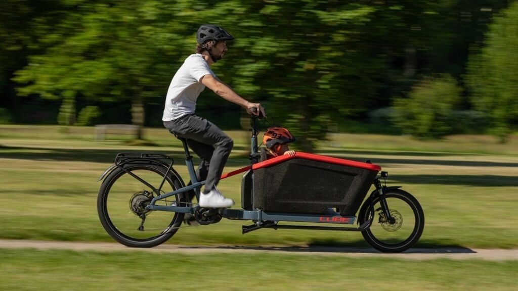 Radfahrer fährt mit Kind im Lastenrad durch einen Park.