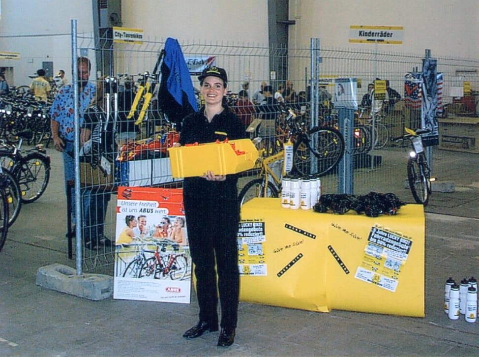 Mitarbeiterin steht mit einem gelben Präsentationskarton vor einem Aktionsstand von Lucky Bike.