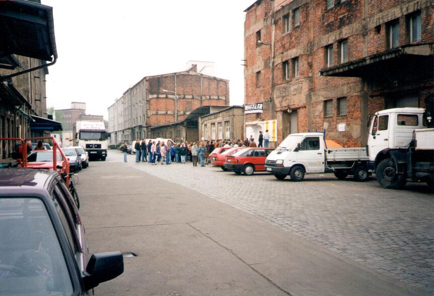 Historische Aufnahme mit einer Menschenschlange und geparkten Autos vor einem Industriegebäude.
