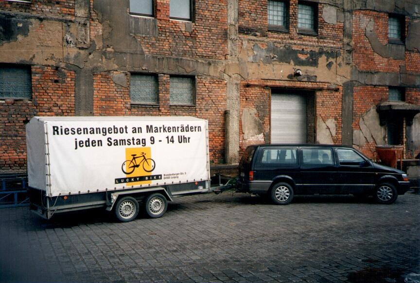 Auto mit Lucky Bike Anhänger vor einem alten Backsteingebäude.