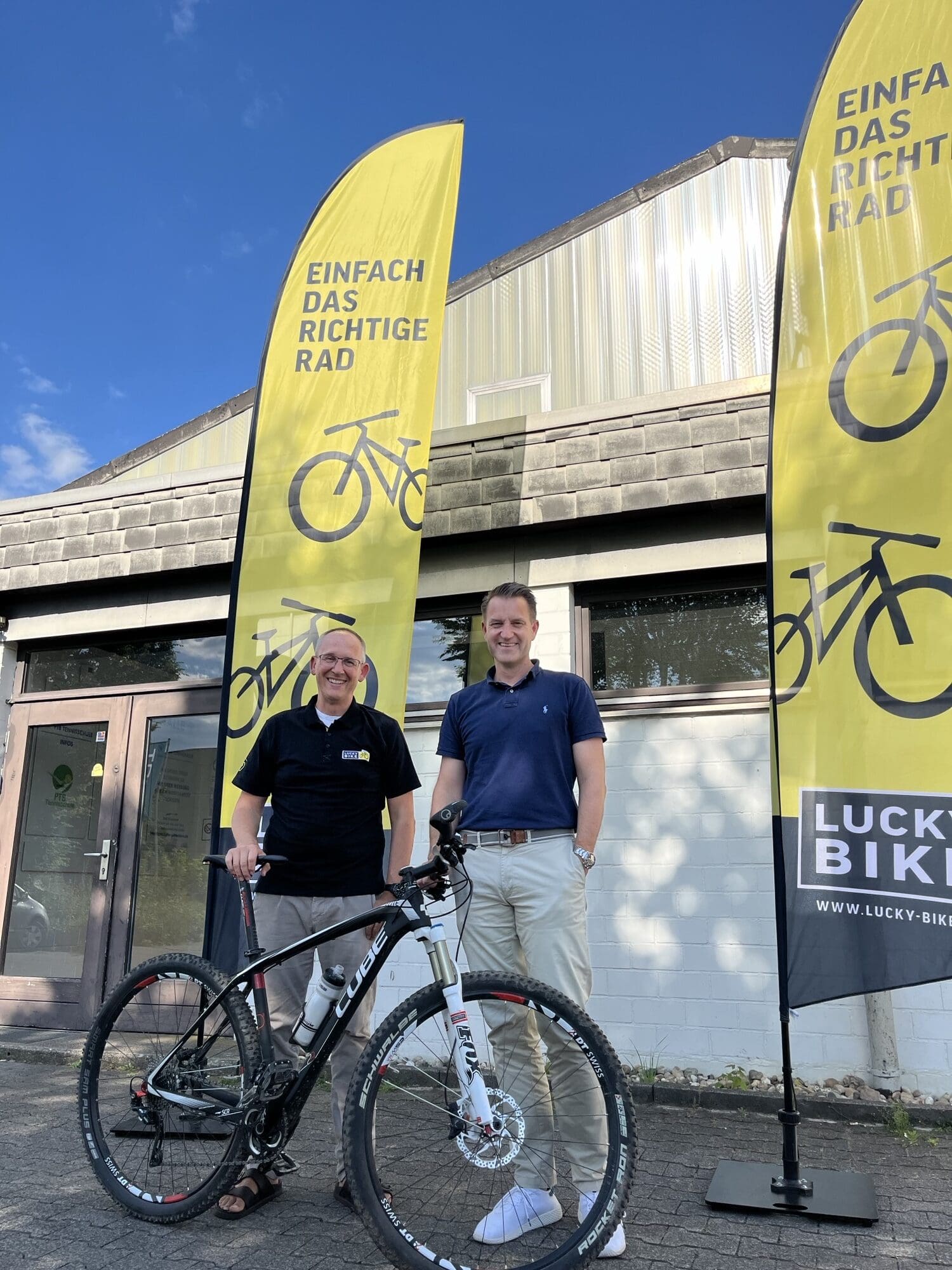 Lucky Bike Geschäftsführer Christian Morgenroth und Architekt Jan-Eric Spork stehen mit einem Mountainbike vor dem Lucky Bike Standort mit gelben Fahnen.