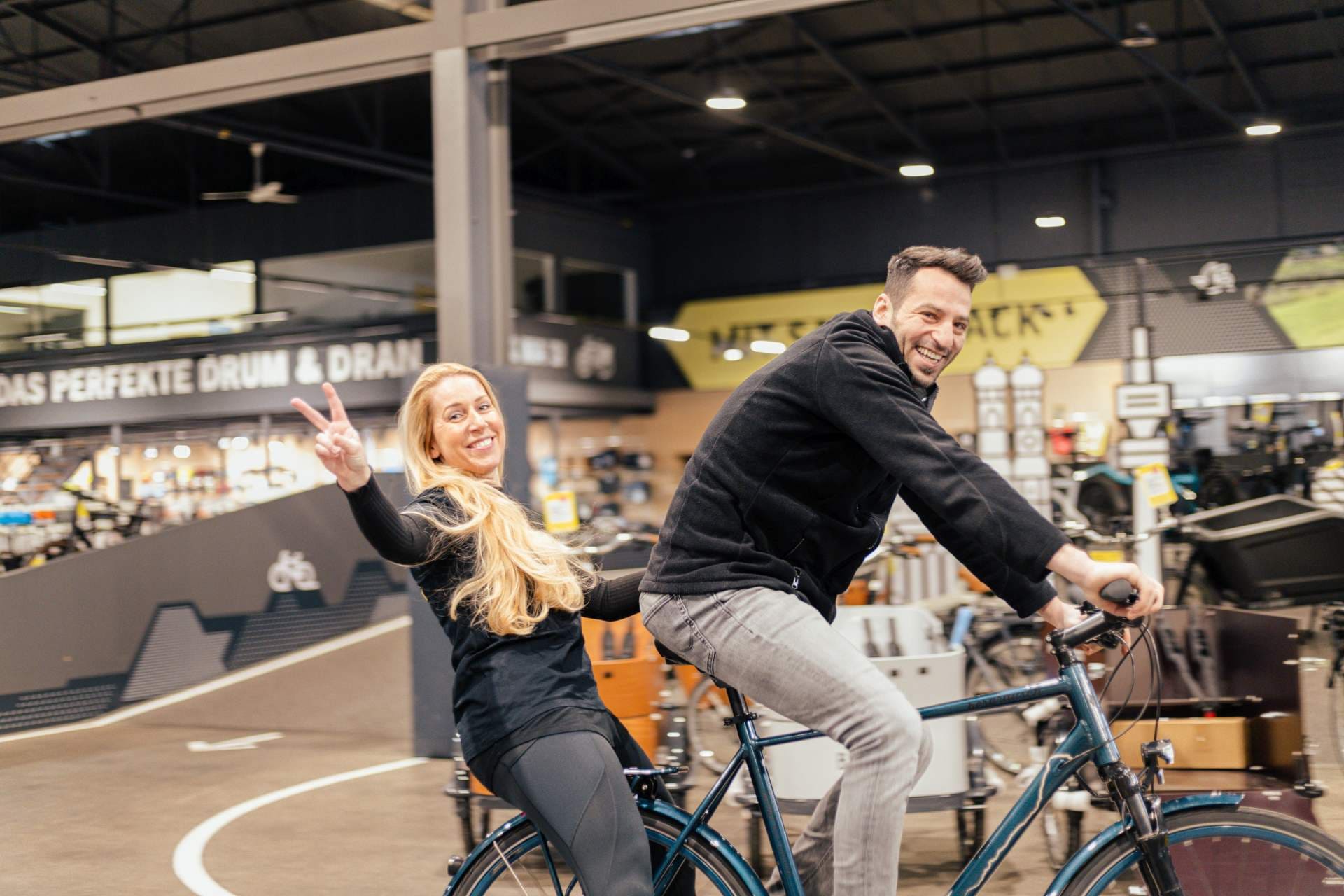 Mann und Frau fahren lachend auf einem Fahrrad über die Verkaufsfläche eines Lucky Bike Geschäfts.