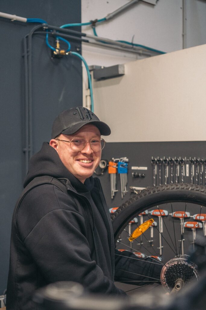 Werkstattleiter montiert Fahrradreifen in moderner Werkstatt, umgeben von Werkzeugen und lächelt in die Kamera.