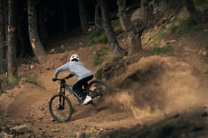 Eine Person fährt mit einem Mountainbike eine Piste entlang und wirbelt dabei mit dem Hinterrad sehr viel Dreck auf